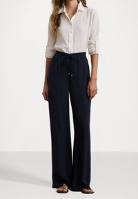 Mujer con una camisa blanca de botones y mangas arremangadas, pantalones anchos de tiro alto en color azul marino con cordón y sandalias marrones, de pie contra un fondo blanco.