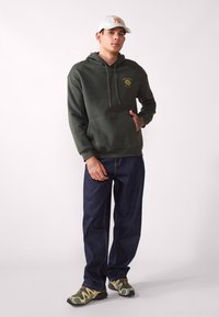 Olivengrønn hettegenser med lomme foran, sammen med mørkeblå jeans og camouflage-sneakers. Aktsom logo på brystet.
