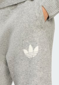 Pantalones de chándal de forro polar gris con cintura elástica acanalada, bolsillos laterales y un logotipo blanco con tres rayas y diseño de hoja en el muslo izquierdo.