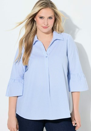 Nuori nainen, jolla on pitkät vaaleat hiukset ja joka käyttää vaaleansinivalkoraidallista paitapuseroa liehuvilla kolmen neljänneksen hihoilla.