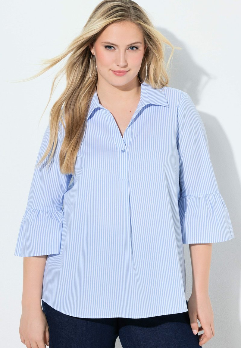 Giovane donna con lunghi capelli biondi che indossa una camicetta a righe azzurre e bianche con maniche a campana a tre quarti.