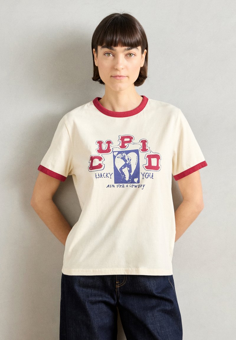 Κρέμα κοντομάνικη μπλούζα με κόκκινη ρίγα στο λαιμό, με εκτυπωμένο γραφιστικό σχέδιο που περιλαμβάνει το κείμενο "Cupid Lucky You" και "Alta for a Cowboy."