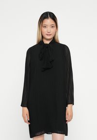 Μαύρο πλισέ φόρεμα με ψηλό λαιμό και λεπτομέρεια φιόγκου, διαθέτοντας μακριά διαφανή μανίκια και άνετη εφαρμογή, που πέφτει πάνω από τα γόνατα.