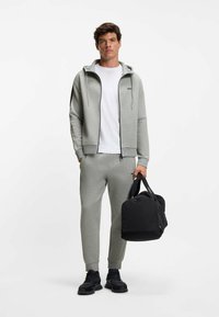 Grijze sweatshirt en joggers, voorzien van een ritssluiting aan de voorkant, capuchon en contrasterende zijstrepen, gecombineerd met een zwarte duffeltas. Geweven stof.