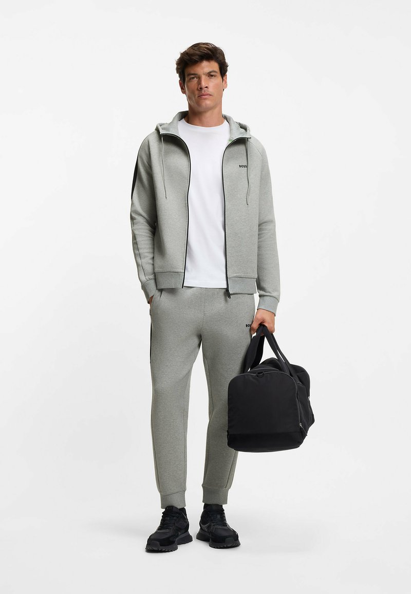 Grijze sweatshirt en joggers, voorzien van een ritssluiting aan de voorkant, capuchon en contrasterende zijstrepen, gecombineerd met een zwarte duffeltas. Geweven stof.