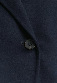 Navy wollen blend jas met een geknipt kraag en een enkele zwarte knop met vier gaten, gemaakt van een zachte, textuur rijke stof.