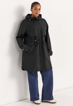 Frau trägt einen schwarzen Kapuzenregenmantel mit großen Fronttaschen, blaue Jeans mit weitem Bein und weiße Sneakers, steht vor einer weißen Wand.