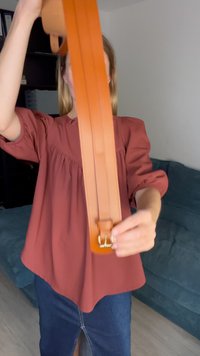 Φαρδιά δερμάτινη ζώνη σε ανοιχτό πορτοκαλί με δύο παράλληλες ραφές, διαθέτοντας χρυσό μεταλλικό κούμπωμα. Εστιασμένη μπροστά σε έναν μπλε καναπέ.