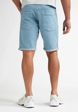 Man draagt lichtblauwe opgerolde korte broek en witte sneakers, gezien van achteren tegen een effen witte achtergrond.