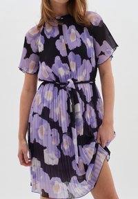 Lila kukallinen laskostettu mekko, jossa on lyhyet hihat, vyöllinen vyötärö ja hieman levenevä hame. Mekossa on rohkeita mustia yksityiskohtia ja pehmeä tekstuuri.