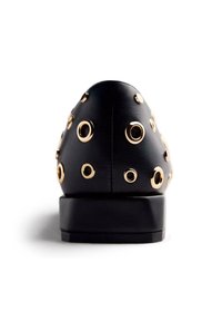 Zapato de cuero negro con punta afilada, adornado con ojales de metal dorado, con suela plana y superficie texturizada.