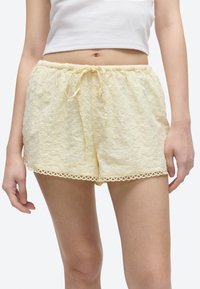 Sieviete valkā gaiši dzeltenas ziedu izšuvumu mežģīņu šorti ar auklas jostu un viļņotu apmali, kombinēti ar īsu baltu tanktopu.