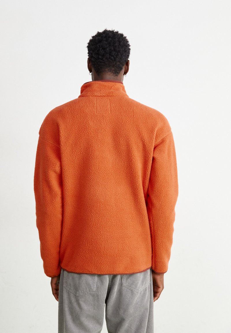 Pullover in fleece arancione con colletto alto, finitura testurizzata e vestibilità comoda. Presenta spalle scese e un'etichetta sul retro.