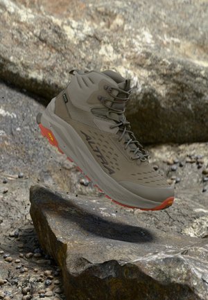 Žygiavimo batai su Vibram padu ir Altra ženklu, pakibę ore virš uolėto reljefo su mažais išsibarsčiusiais akmenėliais ir dideliu akmeniniu fonu.