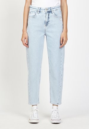 Woman wearing light blue high-waist textured jeans, white sneakers, and a tucked-in white top, standing against a plain background.