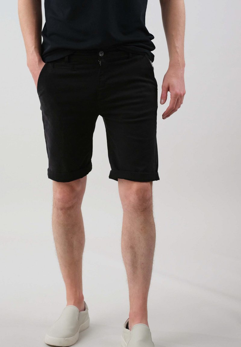 Shorts en coton noir avec des poignets roulés, présentant une fermeture à bouton à l'avant et des poches latérales, associés à des chaussures légères à enfiler.
