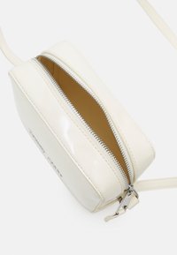 Borsa a tracolla in pelle verniciata bianca con chiusura zip, interno in tonalità nocciola e hardware in argento. Presenta una texture liscia e una forma rettangolare compatta.