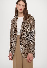 Blazer à imprimé léopard en tan et noir, avec une finition satinée, des revers crantés, deux poches avant et trois boutons. Texture lisse.