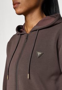 Φόρμα με κουκούλα και φερμουάρ από σκούρο καφέ ύφασμα, με τριγωνική ετικέτα λογότυπου και γυαλιστερά άκρα κορδονιών. Λείος υφαντός ιστός με εφαρμοστή σχεδίαση.