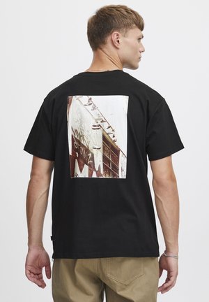 T-shirt en coton noir avec un imprimé graphique rectangulaire au dos, représentant une scène urbaine texturée avec des graffitis et des détails architecturaux.