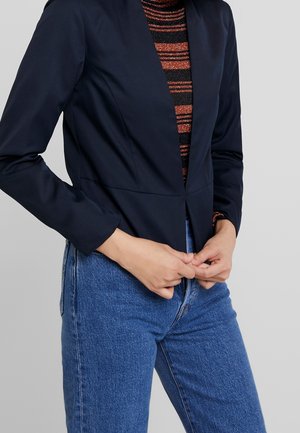 Woman wearing a fitted navy blazer over a black and orange striped top paired with blue jeans, hands clasped at waist.