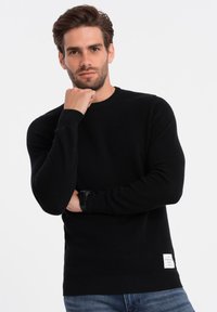 Schwarzer Strickpullover mit langen Ärmeln, gerippten Bündchen, rundem Halsausschnitt und einem gewebten Etikett am unteren Saum. Getragen mit blauer Jeans.