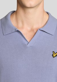 Κοντινό πλάνο του λαιμού και του πάνω μέρους του στήθους με φόρεμα πουλόβερ ανοιχτού μωβ χρώματος με γιακά, με κίτρινο λογότυπο πουλιού στη δεξιά πλευρά.