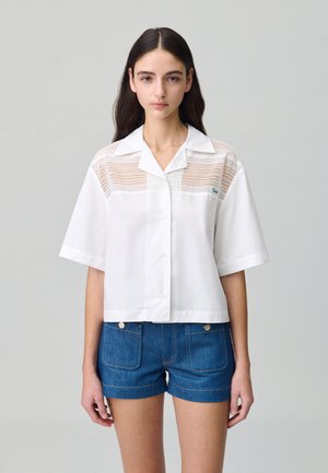 Camicetta bianca a maniche corte con colletto, con pannelli in pizzo sulla parte superiore davanti. Abbinata a pantaloni shorts in denim blu.