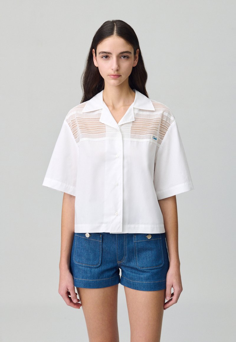 Camicetta bianca a maniche corte con colletto, con pannelli in pizzo sulla parte superiore davanti. Abbinata a pantaloni shorts in denim blu.