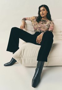 Bluson mit floralen Mustern und langen Ärmeln, schwarze weite Hose und spitze schwarze Stiefeletten, auf einem gerippten, cremefarbenen Sofa sitzend.