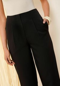 Pantaloni neri su misura con pieghe frontali e tasche laterali. Realizzati in tessuto morbido, presentano una vita aderente e un design a gamba larga.