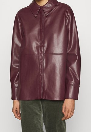 Persoon die een bordeauxrode lange mouwen neplederen blouse draagt met een spitse kraag en verborgen knopen aan de voorkant over olijfgroene broek.
