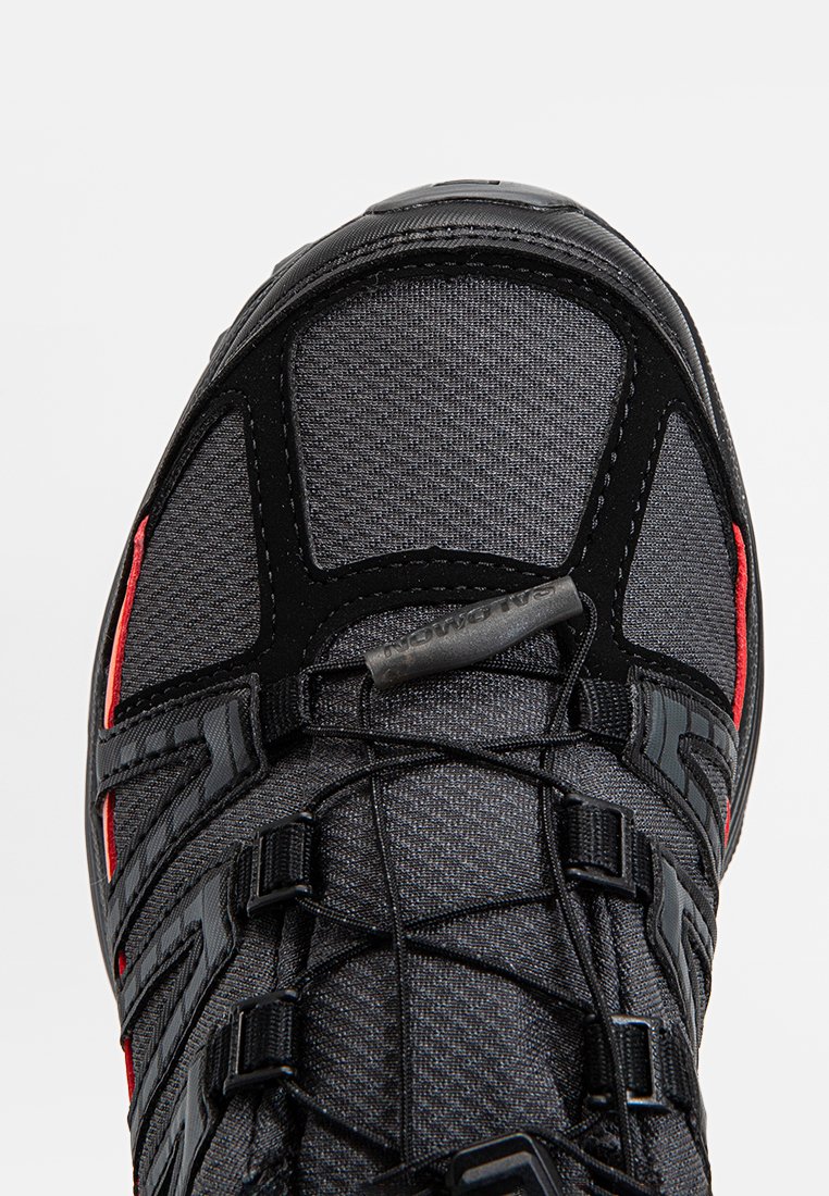 Bout de chaussure de sport noir et gris foncé avec des lacets élastiques noirs et des accents rouges, vu de dessus sur un fond blanc.