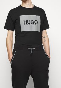 Schwarzes T-Shirt mit einem grauen rechteckigen Logo, das "HUGO" und "HUGO BOSS" zeigt. Begleitet von schwarzen Jogginghosen mit Seitentaschen und Kordelzug.