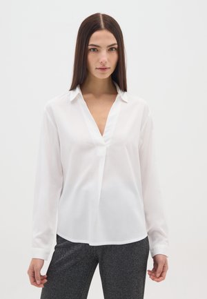 Mujer con blusa blanca de manga larga y escote en V y pantalones gris oscuro, posando frente a un fondo liso y claro.