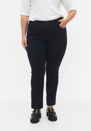 Jeans ajustados de color azul oscuro con una textura suave, diseño de cinco bolsillos y costuras en contraste. Combinados con mocasines negros y una blusa blanca.