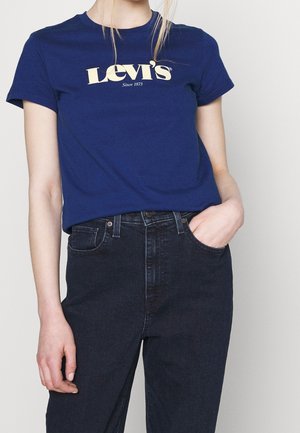 Person wearing a blue Levi's t-shirt and black high-waist jeans, with one hand in pocket and light background.