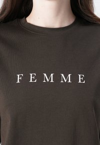 Donkerbruine katoen t-shirt met een gecentreerde witte opdruk met de tekst "FEMME." Klassieke ronde hals en zachte textuur. Eenvoudig ontwerp zonder patronen.