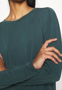 Långärmad skjorta i teal med rund halsringning, slät textur och subtila axelsömmer som accent, med korsade armar som vilar mot kroppen.