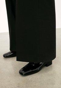 Pantofi din piele patent neagră, cu vârf pătrat și toc mic bloc, purtați cu pantaloni largi negri, expuși pe o suprafață de beton.