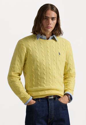 Geel kabelgebreide trui met een ronde halslijn, gedragen over een lichtblauw overhemd met kraag. Gekleed met donkere spijkerbroek. Casual ontwerp.