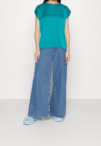 Top in satin color teal con maniche corte abbinato a jeans in denim leggeri a gamba larga di colore blu, completato da sandali platform blu chunky.