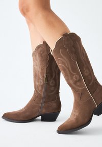 Botas de vaquero de ante marrón con puntas afiladas, costuras decorativas y una cremallera lateral, que presentan un tacón bloque acampanado y un ribete a juego.