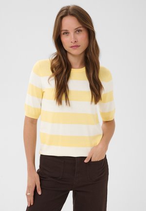 Ung kvinde med langt brunt hår iført en gul og hvid stribet kortærmet sweater og mørke bukser med den ene hånd i lommen.