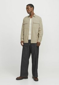 Camicia a righe beige con bottoni e due tasche sul petto, indossata sopra un tank top crema; abbinata a jeans larghi scuri e stivali marroni.