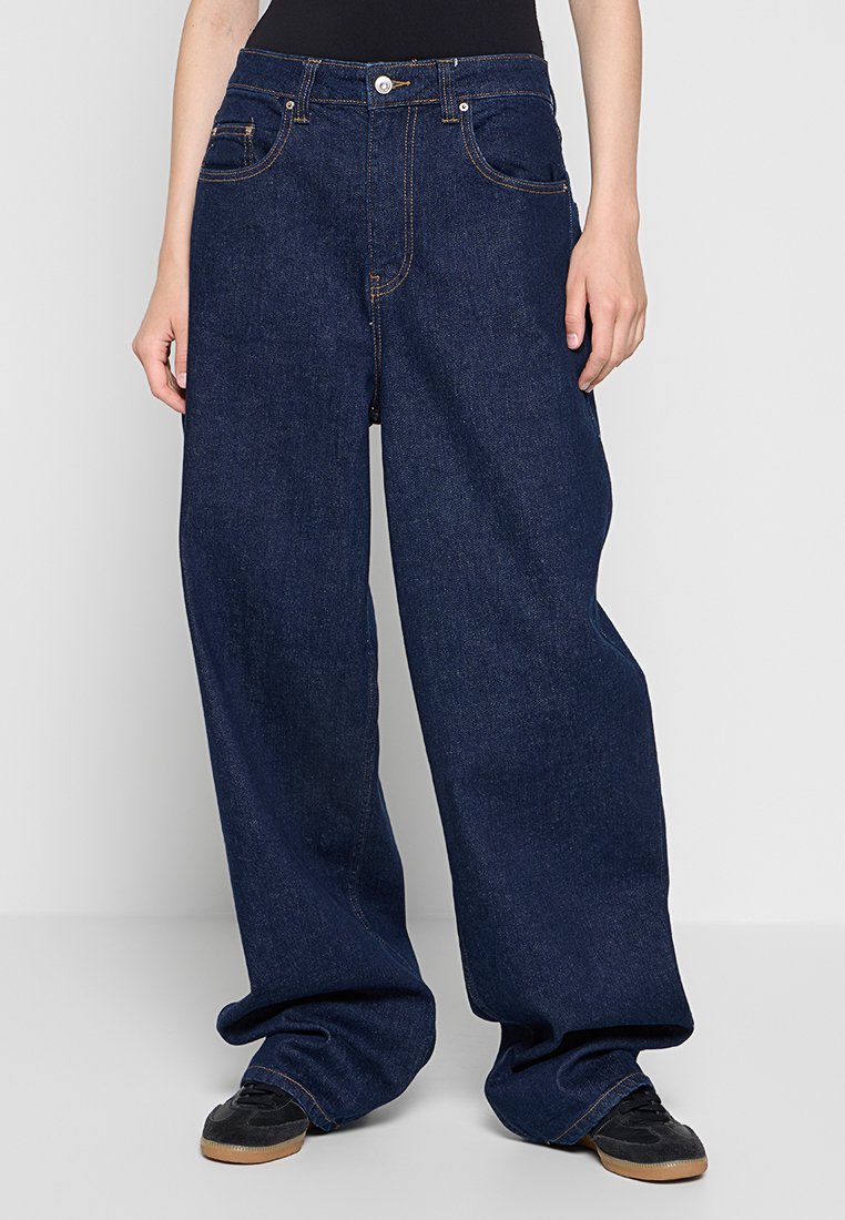 Jeans in denim a gamba larga di colore blu scuro con cuciture a contrasto, dotati di tasche frontali e chiusura con zip. Tessuto morbido e resistente con vestibilità comoda.