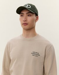 Gorra caqui con logo blanco y ajuste en la parte posterior; sudadera beige con "LES DEUX COPENHAGEN EST. 2011" bordado en el pecho.