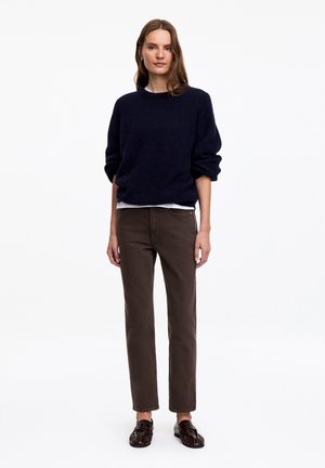Femme debout portant un pull bleu foncé, un maillot blanc, un pantalon marron et des mocassins marron foncé sur un fond blanc uni.