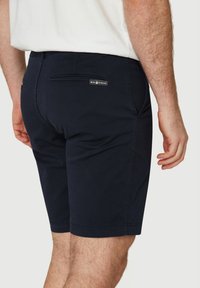 Tummansiniset shortsit, jotka on valmistettu sileästä kankaasta. Niissä on takatasku, jossa on brändilogo, ja ne on suunniteltu siisteillä linjoilla ilman näkyviä kuvioita.