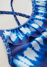 Blauwe zwemkleding met een gerimpelde halslijn, een tie-dye patroon in tinten blauw en wit, en kruislings gesneden bandjes met een getextureerde rand.